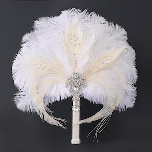 May include: A bridal bouquet featuring white and cream-colored feathers, accented with sparkling rhinestones and a silver-toned, ornate centerpiece. The handle is wrapped in a cream-colored ribbon.
