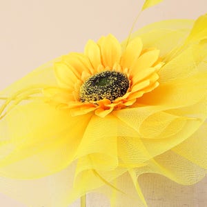 May include: A vibrant yellow headpiece with a large sunflower and a dark green centre. The sunflower is surrounded by layers of yellow netting and accented with delicate yellow feathers, creating a cheerful and elegant design.