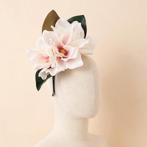 Peut inclure: Un accessoire de tête floral avec de grandes fleurs roses pâles et blanches, avec des centres jaunes, accentuées par des feuilles vert foncé. Les fleurs sont fixées sur un serre-tête noir.