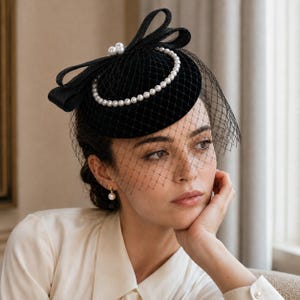 May include: A black fascinator hat featuring a circular base, a large structured bow, and a delicate black netting veil. The hat is adorned with a circular arrangement of white faux pearls and a cluster of three larger pearls at the top of the bow.
