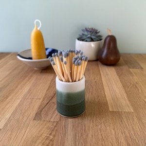May include: A ceramic match holder with a white and green ombre design, filled with matches with grey tips. A yellow candle, a small succulent, and a pear-shaped object are in the background.