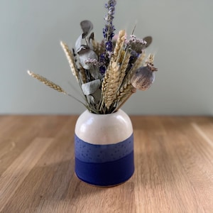 May include: A small ceramic vase with a white top, a blue speckled middle section, and a solid blue base. The vase contains a dried flower arrangement with wheat, lavender, and seed pods. The vase sits on a wooden surface.