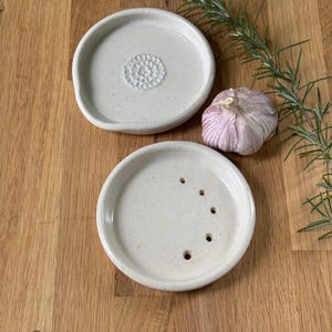 May include: Two white ceramic garlic crushers. One has a textured surface and the other has holes for pressing garlic. A clove of garlic and a sprig of rosemary are on a wooden surface.