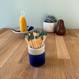 May include: A ceramic match holder with a blue base and cream top, filled with wooden matches with green tips. A yellow candle, a succulent in a white pot, and a wooden pear are also on the wooden surface.