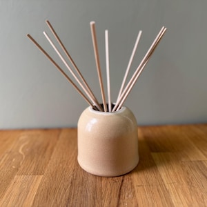 May include: A beige ceramic diffuser bottle with a round top and a white rim. The bottle is filled with wooden diffuser sticks.