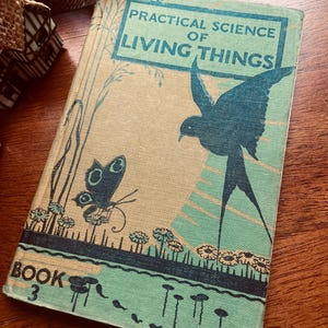 May include: Vintage book titled "Practical Science of Living Things, Book 3." The cover features a teal and beige illustration of a bird, butterfly, and flowers. The book has a worn appearance, suggesting it is an antique.