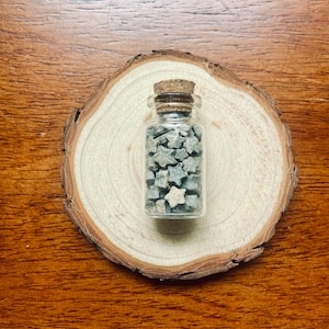 Könnte beinhalten: Eine kleine Glasflasche gefüllt mit grauen, sternförmigen Steinen, verschlossen mit einem Korken. Die Flasche steht auf einer runden, hellen Holzscheibe, vor einem dunklen Holzgrund. Die Sterne sind etwa 1,3 cm groß.