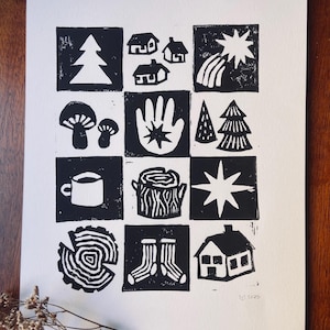 Op de afbeelding: Zwart-wit linoleumsnede met een raster van illustraties met een natuurthema. De prent toont afbeeldingen van bomen, huizen, paddenstoelen, een hand met een ster, een mok, een boomstronk, een ster, een boomring, sokken en een huis. De prent staat op wit papier.