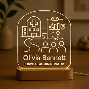 May include: A personalized LED night light with a wooden base. The clear acrylic panel features a hospital-themed design with a building, medical chart, and stethoscope. The name "Olivia Bennett" and title "HOSPITAL ADMINISTRATOR" are displayed.