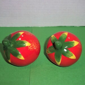 May include: Two ceramic tomato salt and pepper shakers. The shakers are red with green and yellow details. The shakers are on a green surface.