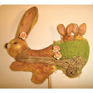 May include: A brown bunny with pink flowers on its head pulling a green wagon with two smaller brown bunnies inside. The wagon is made of fabric and has a brown rope tied around it.
