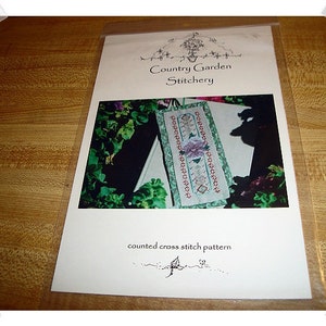 May include: A white cross stitch pattern with a floral design and the text "Country Garden Stitchery" and "counted cross stitch pattern".