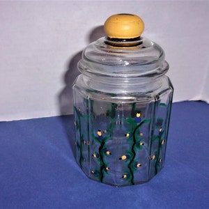 May include: A clear glass jar with a lid and a yellow knob. The jar has an octagonal shape and is decorated with green vines, blue flowers, and yellow accents. The jar is likely used for storage.