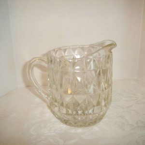 May include: A clear glass creamer with a diamond pattern. The creamer has a handle and a spout.