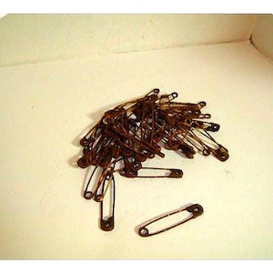 May include: A pile of rusty safety pins, in various sizes, scattered on a light-colored surface. The pins are a dark, aged brown color, suggesting they are vintage or have been exposed to the elements. The pins are open and closed.