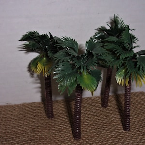 May include: Four miniature plastic palm trees with brown trunks and green fronds. The trees are arranged on a brown burlap surface.