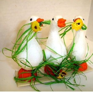 May include: Three handmade white goose figurines with orange beaks and feet, each holding green grass and a yellow flower. The geese are arranged on a light-colored surface, creating a whimsical decorative display.