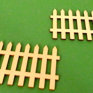 May include: Two small, unfinished wooden picket fences on a green background. The fences are light brown and have pointed tops. The image is well-lit and the fences are in sharp focus.