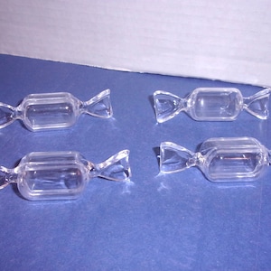 May include: Four clear glass candy-shaped dishes. Each dish has a rectangular center with twisted ends, resembling wrapped candies. The dishes are transparent and sit on a blue surface.