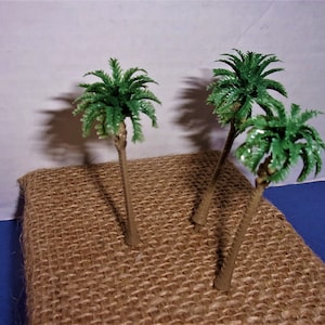 May include: Three miniature plastic palm trees with green fronds on a brown burlap surface.