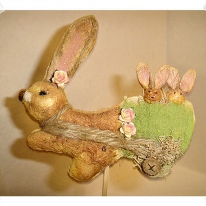 May include: A charming Easter decoration features a tan bunny crafted from textured paper mache.  The bunny is adorned with small pink flowers and carries two smaller bunnies in a green, moss-like cart secured with twine. The cart is accented with additional pink flowers and straw-like elements. This whimsical piece is perfect for spring decor or as a unique Easter gift.