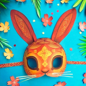 Puede incluir: Una máscara decorativa de conejo de papel con un vibrante esquema de color naranja y rojo. La máscara presenta intrincados patrones dorados y largas orejas erguidas. El fondo es azul brillante, adornado con flores y hojas de papel.
