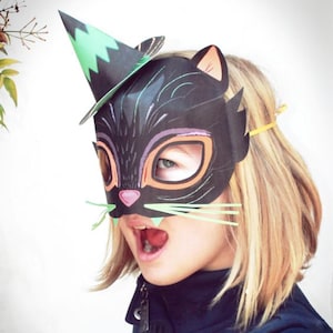May include: A black cat mask with green and orange accents. The mask has a pointy hat on top and a pink nose. The mask is being worn by a child with blonde hair.
