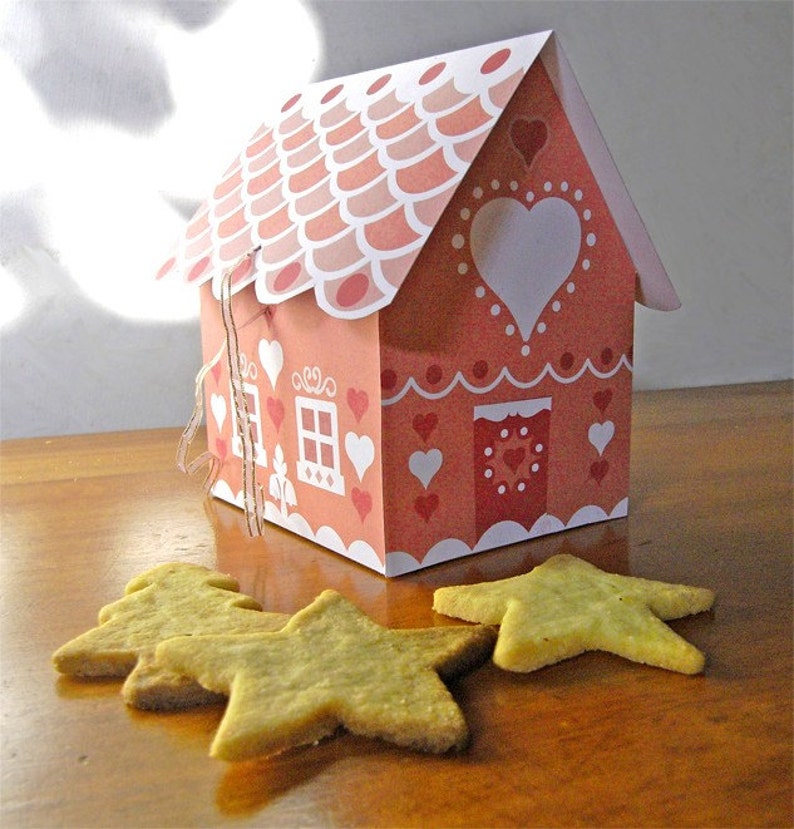 May include: A pink and white gingerbread house with a white heart on the front and a red door. The house is decorated with white dots and hearts. There are four star-shaped cookies on a wooden surface in front of the house.