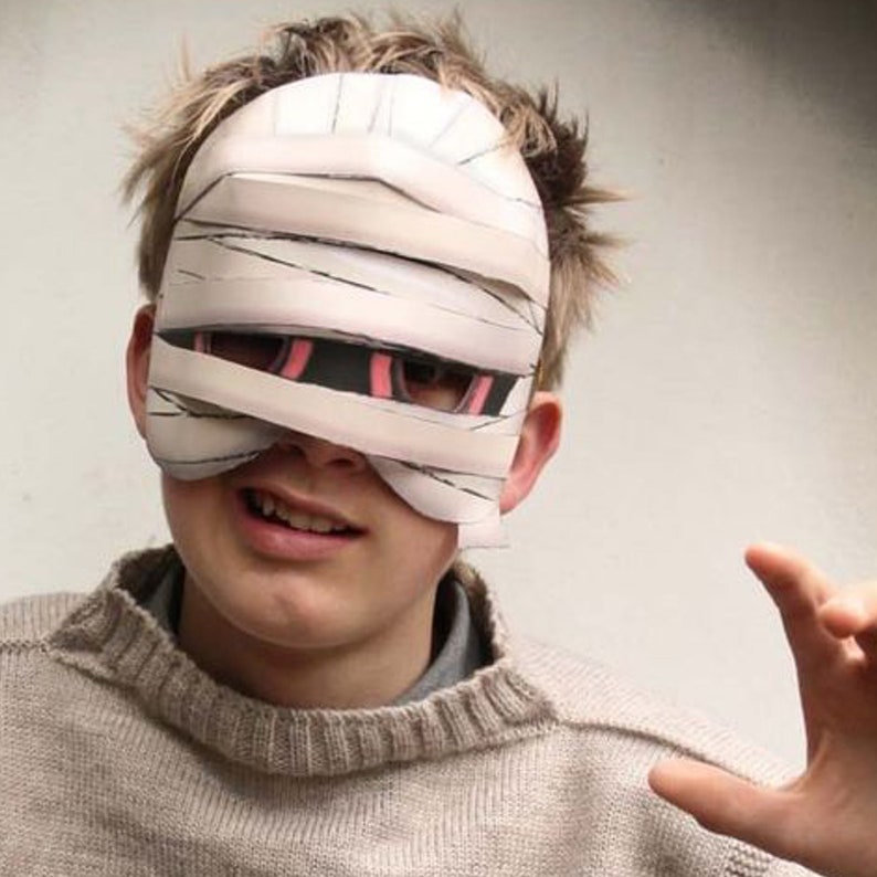 May include: A white paper mask with black and pink eye holes, resembling a mummy bandage. The mask is worn by a person with short brown hair.