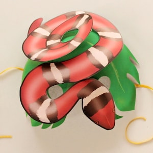 May include: A red and white snake mask with a green leaf design. The mask has yellow string ties.