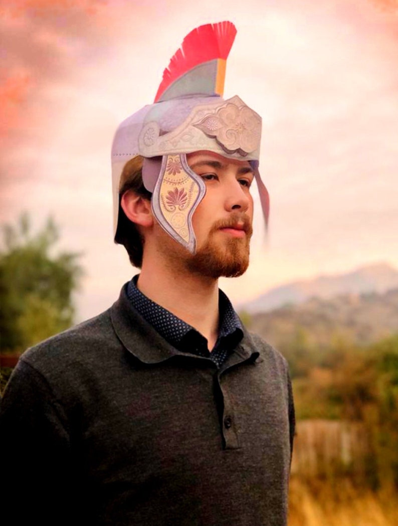 May include: A person wearing a gray sweater and a paper helmet with a red, green, and white plume. The helmet has a decorative design with a cloud and floral patterns.