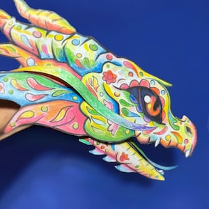 Könnte beinhalten: Ein mehrfarbiger Papierdrachenkopf-Ausschnitt mit komplizierten handgezeichneten Mustern in Gelb, Grün, Blau, Pink und Orange. Das Design zeigt florale Motive, wirbelnde Formen und ein großes dunkles Auge mit einer leuchtend orangefarbenen Iris.