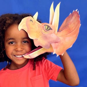 May include: A young person wearing a red shirt holds a large orange and yellow paper dinosaur mask up to their face. The mask is a triceratops with a large mouth and three horns.