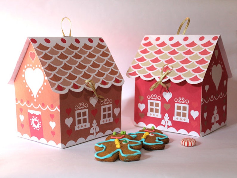 May include: Two gingerbread house shaped boxes with red and brown accents. The boxes have white hearts and windows. The boxes are decorated with gold ribbon. There are gingerbread cookies in front of the boxes.