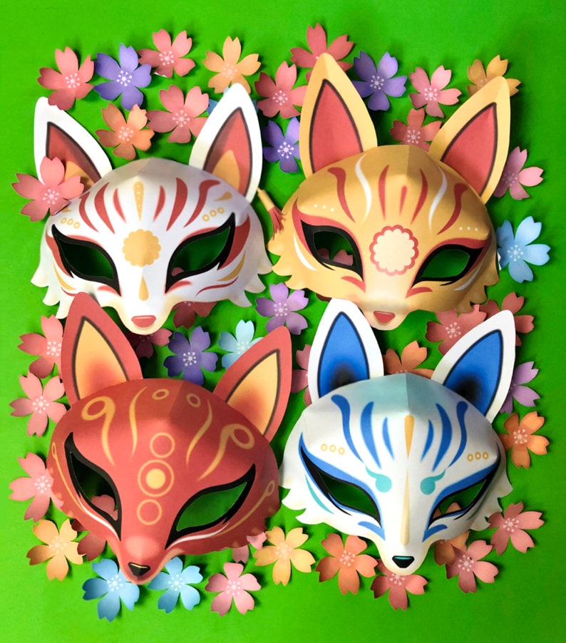 Puede incluir: Cuatro m&aacute;scaras japonesas kitsune de colores con dise&ntilde;os intrincados, sobre un fondo verde con flores de papel rosa y azul.