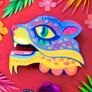Puede incluir: Una máscara de jaguar de papel maché colorida con detalles azules, amarillos, rosas y verdes. La máscara tiene una boca grande abierta con dientes afilados y un ojo detallado.
