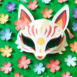 Puede incluir: Una m&aacute;scara de zorro blanca con detalles rojos y dorados, rodeada de flores de papel rosas, azules y amarillas sobre un fondo verde.