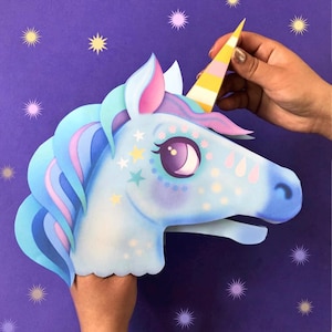 May include: A blue unicorn paper mask with a pink and yellow horn, decorated with stars and glitter. The mask is held by a hand against a purple background.