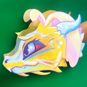 May include: A colorful, layered paper dragon head with yellow, blue, pink, and white accents. The dragon has a detailed eye, horns, and a toothy grin. The artwork is set against a solid green background.