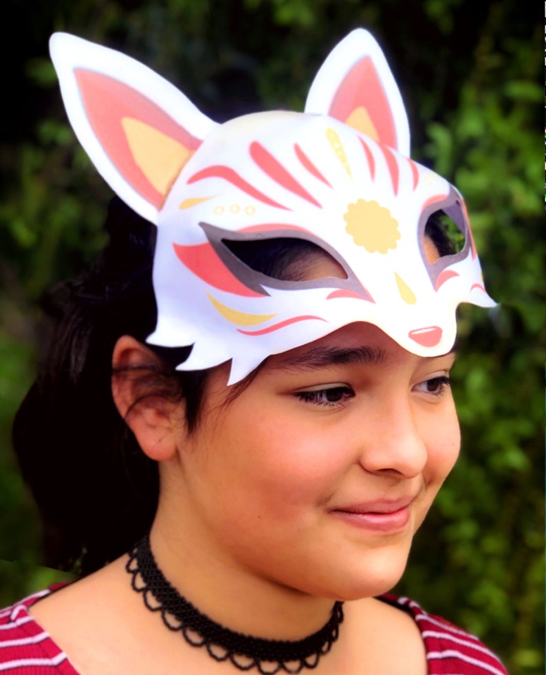 Puede incluir: Una m&aacute;scara de zorro blanca con detalles rojos y dorados. La m&aacute;scara tiene un rostro de zorro estilizado con ojos grandes y una nariz puntiaguda. La m&aacute;scara la lleva puesta una persona con el pelo oscuro.