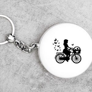 K&ouml;nnte beinhalten: Ein silberner Schl&uuml;sselanh&auml;nger mit einem runden wei&szlig;en Anh&auml;nger, der eine schwarze Silhouette einer Person auf einem Fahrrad mit einem Korb zeigt. Schmetterlinge sind um die Person und das Fahrrad herum verstreut.