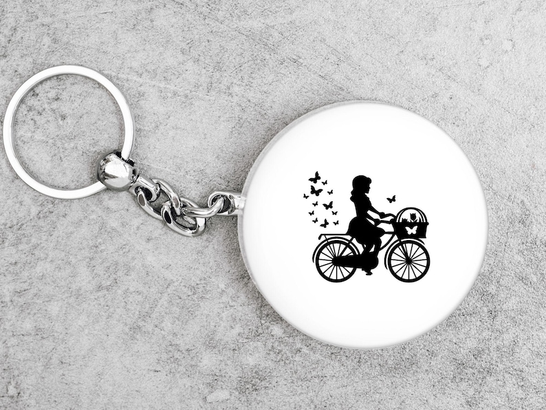 K&ouml;nnte beinhalten: Ein silberner Schl&uuml;sselanh&auml;nger mit einem runden wei&szlig;en Anh&auml;nger, der eine schwarze Silhouette einer Person auf einem Fahrrad mit einem Korb zeigt. Schmetterlinge sind um die Person und das Fahrrad herum verstreut.