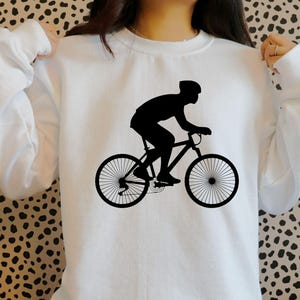 K&ouml;nnte beinhalten: Wei&szlig;es Sweatshirt mit einer schwarzen Silhouette einer Person auf einem Fahrrad.