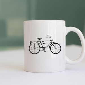 K&ouml;nnte beinhalten: Wei&szlig;e Keramiktasse mit einer schwarzen Silhouette eines Fahrrads mit einer Tasche auf dem Gep&auml;cktr&auml;ger.