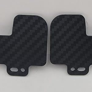 以下が含まれることがあります： 黒いカーボンファイバー製のパーツが2つ。織り目がテクスチャ加工されています。各パーツは丸みを帯びた上部と、2つの小さな穴がある長方形の延長部を持っています。白い背景に表示されています。