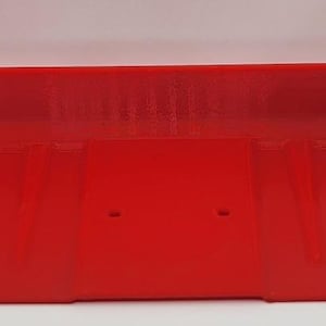 May include: A bright red plastic tray with raised edges and two small holes. The tray has a rectangular shape with curved ends, designed for holding items. The overall design is simple and functional.