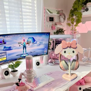 May include: A pink and white Hello Kitty themed headset stand with a heart-shaped base. The stand is on a white desk with a pink and white keyboard, a microphone, and a computer monitor displaying a video game.