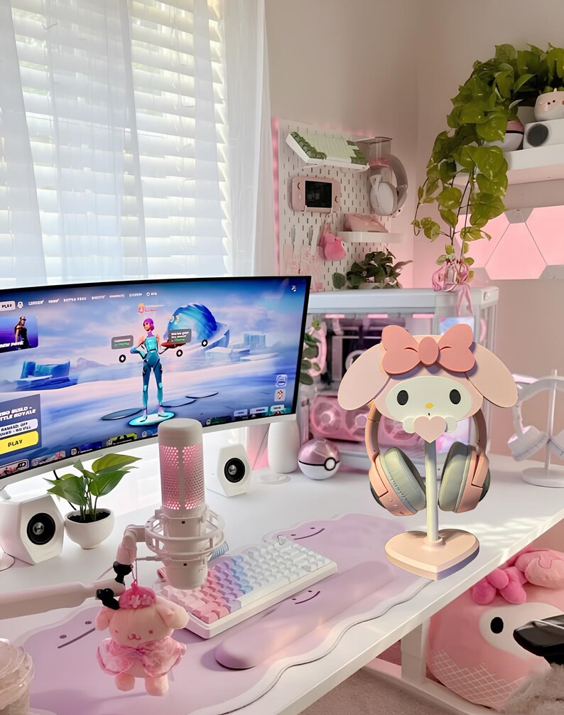 May include: A pink and white Hello Kitty themed headset stand with a heart-shaped base. The stand is on a white desk with a pink and white keyboard, a microphone, and a computer monitor displaying a video game.