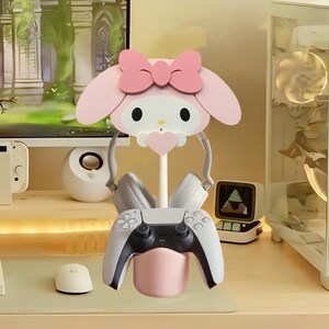 May include: A pink Hello Kitty themed gaming headset stand with a grey controller and headphones. The stand has a pink base and a Hello Kitty head with a pink bow.