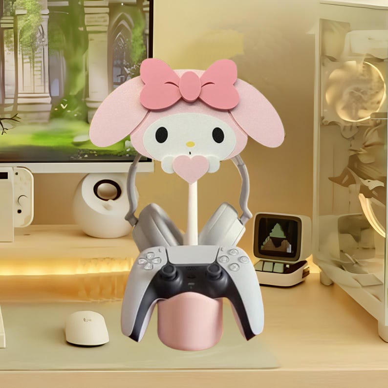 May include: A pink Hello Kitty themed gaming headset stand with a grey controller and headphones. The stand has a pink base and a Hello Kitty head with a pink bow.